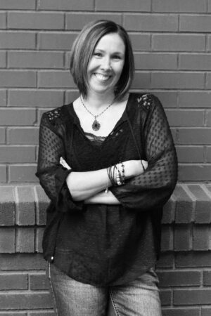 A black and white photo of author, erin Jensen, standing in front of a brick wall. Erin is wearing jeans and a darkers top with spotted mesh sleeves. Her arms are folded, she is wearing jewellery on her right wrist and a neclace round her neck. Erin wears her hair in a long bob style and she is smiling. 