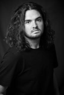 Black and white photo of Josiah Hartley. He has long dark hair wavy hair and is looking at the camera 