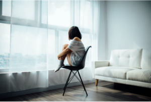 Image of a girl sitting a a chair, knees tucked up, back to the camera facing a net curtain covered window